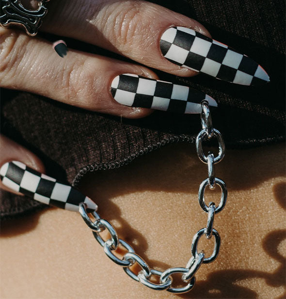 Model wears black and white checkered press-on nails with silver chain attached to pinky and ring fingers