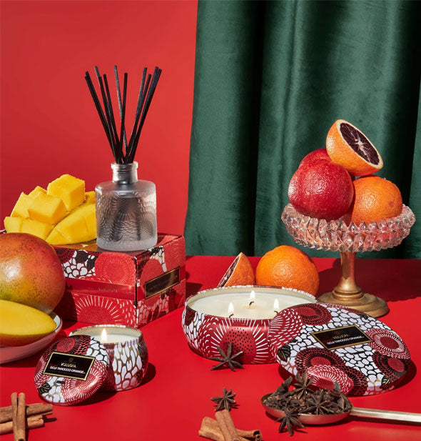 Small and large decorative red and white tin candles and embossed glass reed diffuser are staged with orange fruits, star anise, and cinnamon sticks