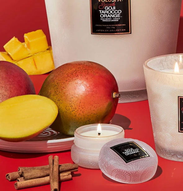 Several Voluspa Spiced Goji Tarocco Orange candles in white embossed glass are staged with mangoes and cinnamon sticks