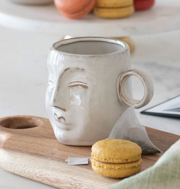 Gray stoneware mug with face shape on the front is staged with teabag and macaron