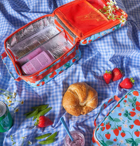 Strawberry print lunch bags staged on a blue gingham cloth with flowers, snap peas, strawberries, a smoothie, and a croissant