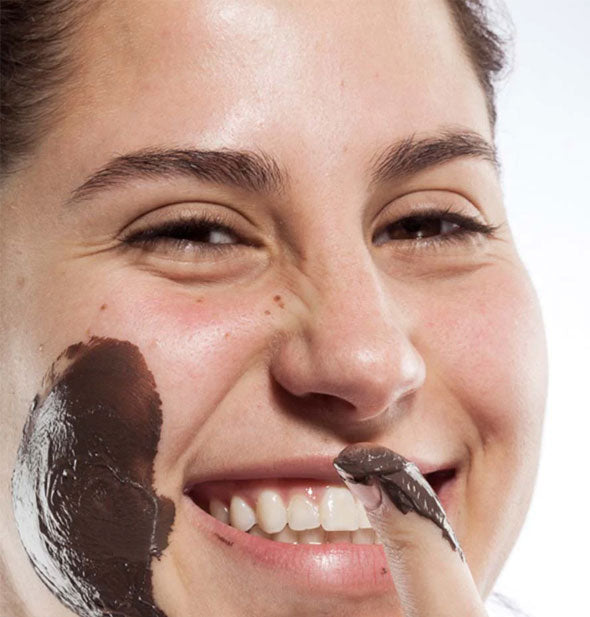 Smiling model with chocolate mask applied to cheek holds out a fingertip covered in mask