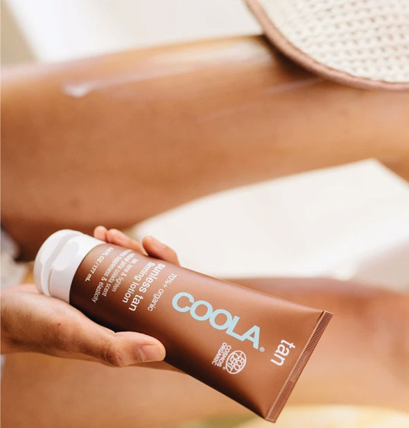 A model holds a bottle of COOLA Tan lotion and applies to leg in background