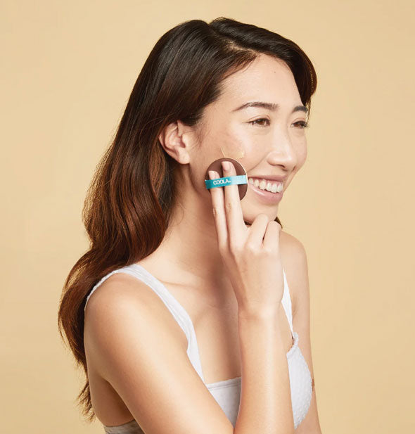 Smiling model applies sunless tanner to cheek with a compact applicator