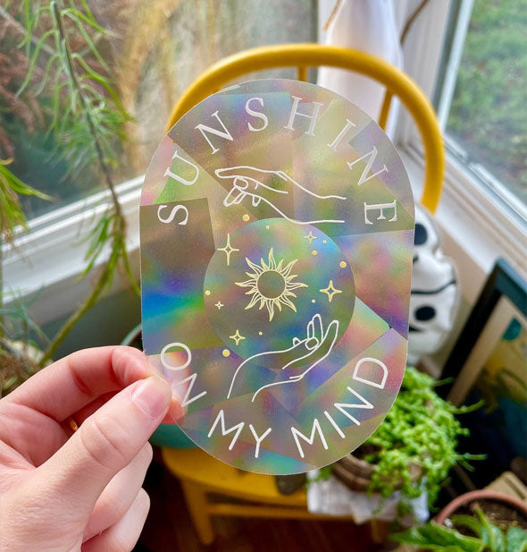 Model's hand holds an oval-shaped "Sunshine on My Mind" sticker with white outlined artwork and a prismatic effect