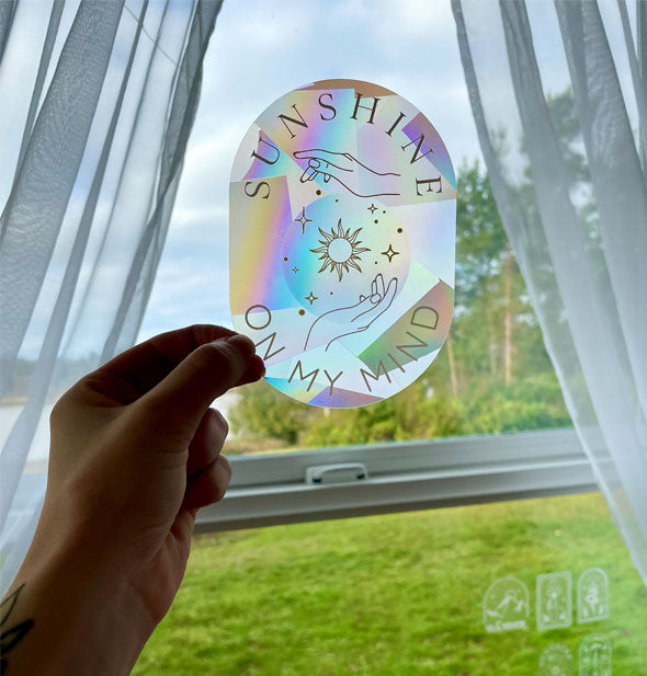 Model's hand holds a prismatic-effect Sunshine on My Mind sticker up in front of a window