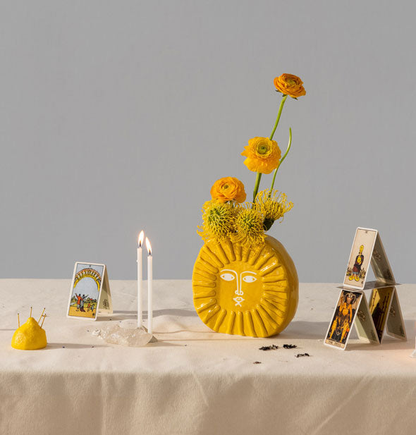 Sun vase with orange wildflowers in it is staged on a tabletop with tarot cards, taper candles, half a lemon, and other items