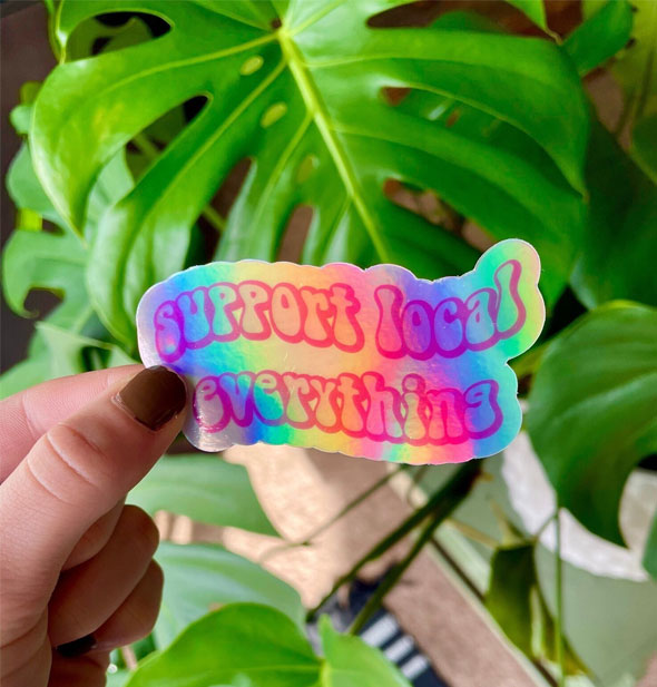Model's hand holds a rainbow holographic sticker that says, "Support local everything" in wavy retro lettering in front of a backdrop of green monstera leaves