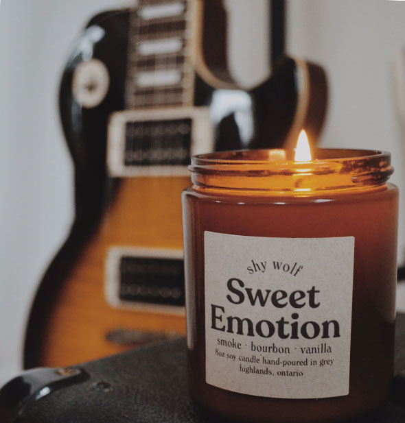 A lit Sweet Emotion candle by Shy Wolf sits on top of an amp with an electric guitar in the background