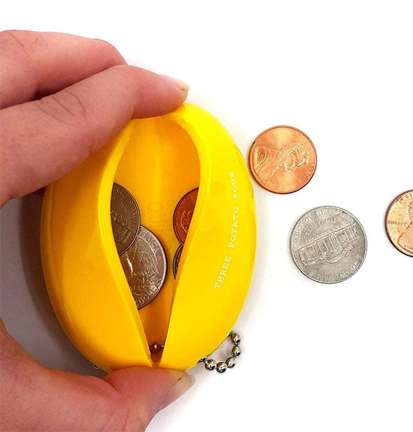 A hand squeezes open a yellow rubber coin pouch to reveal loose change inside