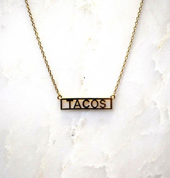 Gold bar necklace on white marble surface says, "Tacos"