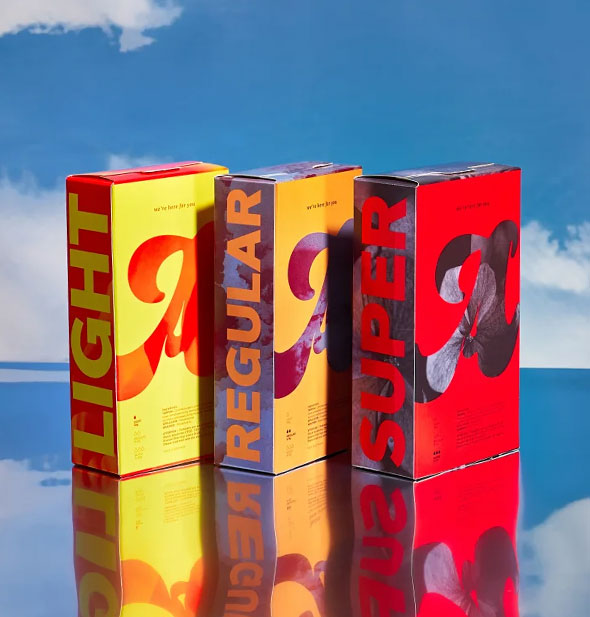 Brightly colored boxes of light, regular, and super absorbency August brand tampons on a background of blue sky with white clouds