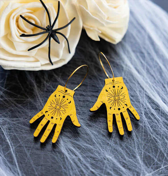 Pair of golden tarot/palmistry hand earrings on a white fibrous surface with white roses and a black plastic spider
