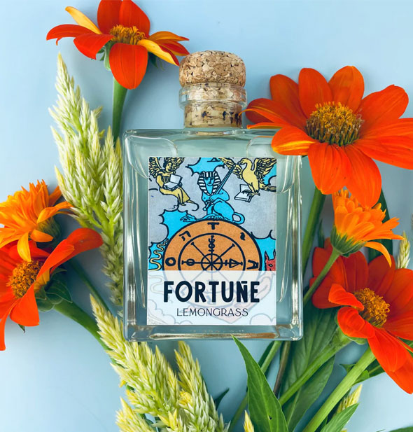 Square glass bottle with Fortune tarot card label and cork lid rests on a blue surface with orange daisies and yellow cockscomb