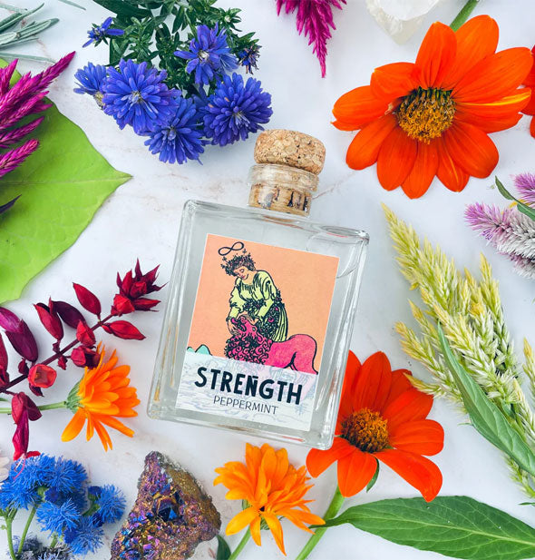 Square glass bottle with Strength tarot card label and cork lid rests among colorful flowers