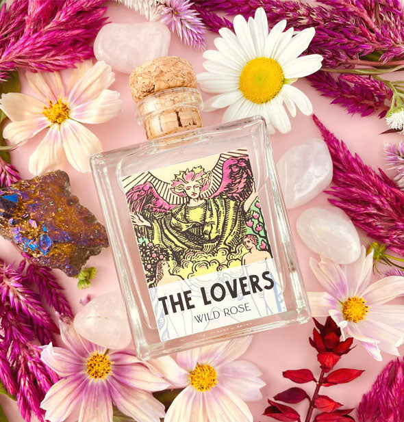 Square glass bottle with The Lovers tarot card label and cork lid rests among purple cockscombs and pink daisies