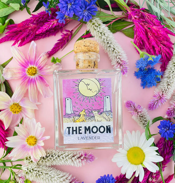 Square glass bottle with The Moon tarot card label and cork lid rests among colorful flowers on a pink surface