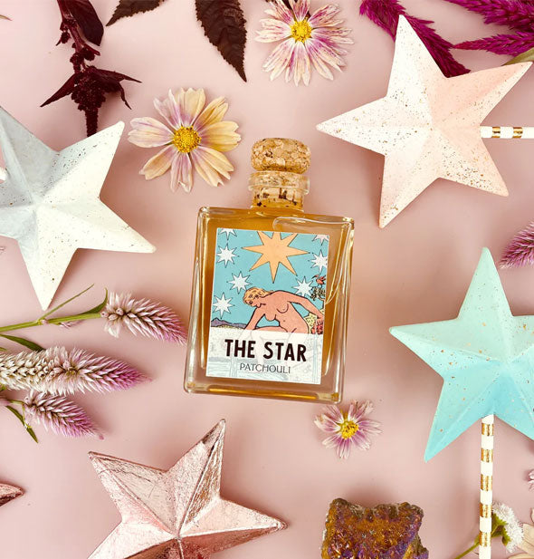 Square glass bottle with The Star tarot card label and cork lid rests on a pink surface with flowers and star wands