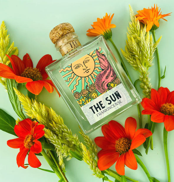 Square glass bottle with The Sun tarot card label and cork lid rests among brightly colored flowers on an aqua surface