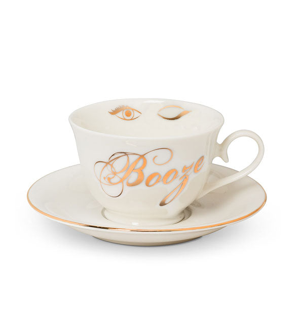 White teacup with "Booze" in metallic gold cursive and winking eye graphic on its inner rim sits on a gold-rimmed saucer