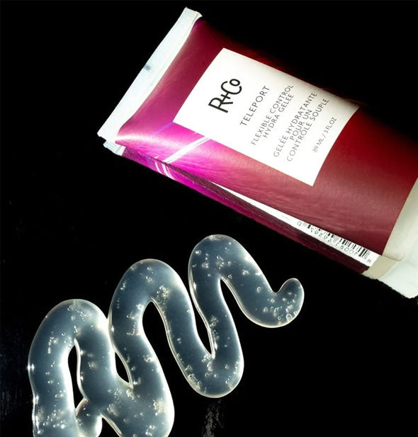 Bottle of R+Co Teleport Flexible Control Hydra Gelee on a mirrored surface next to a squiggled application of product, which is mostly clear with bubbles throughout
