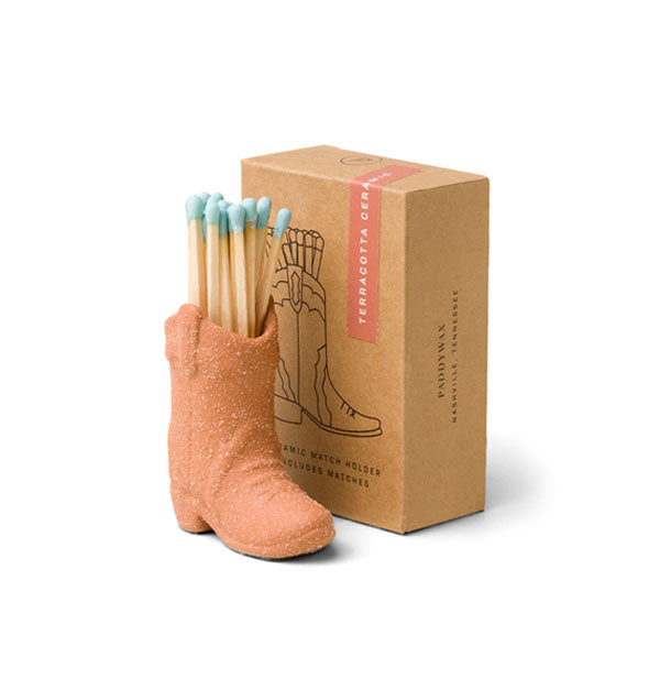 Terracotta cowboy boot match holder with blue-tipped wooden matches inside next to brown box packaging
