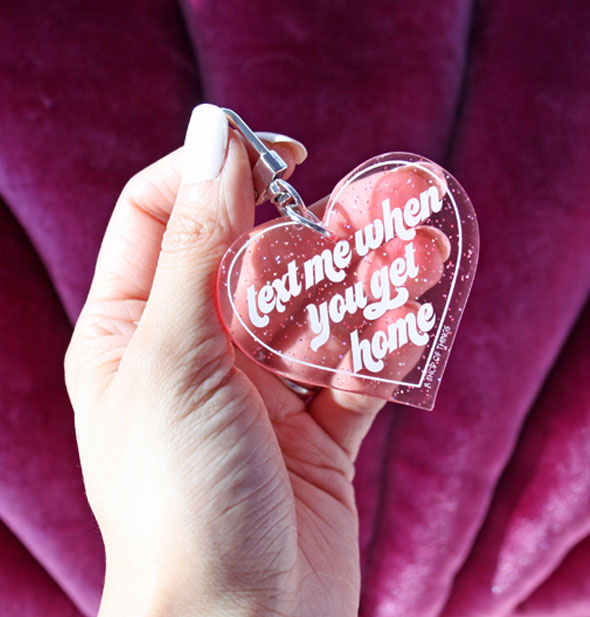 Model's hand holds the Text Me When You Get Home glitter heart keychain in front of a plush pink backdrop