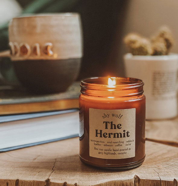 The Hermit amber glass jar candle shown lit on a wooden surface with books and other home goods in the background