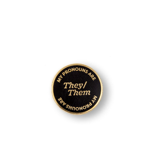 Round black enamel pin with gold border says, "My Pronouns Are" at top and bottom and "They/Them" in the center, all in gold lettering