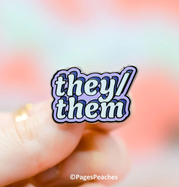 A purple enamel They/Them pin is held between a model's fingers with Pages Peaches image copyright at bottom