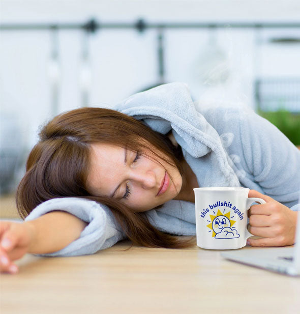Model in blue robe with head resting on arm on a tabletop holds a This Bullshit Again mug