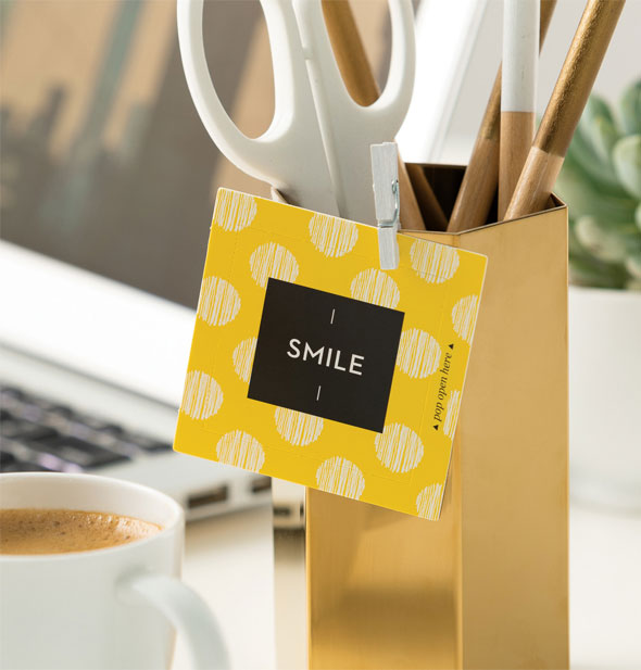 A yellow polka dotted Smile card hangs by a small white clothespin from a pencil cup on an office desktop