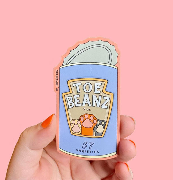 Model's hand holds a sticker shaped like a can with label that says, "Toe Beanz" with image of cat paws