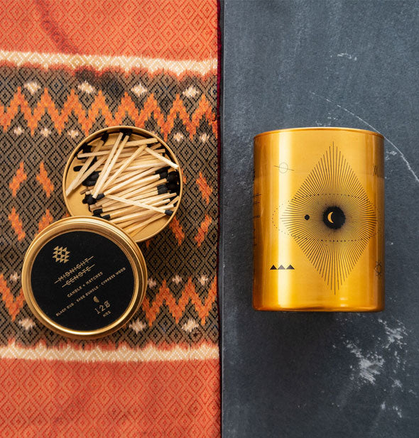 Metallic gold moon candle lays on a stone surface next to an opened tin lid with black-tipped matches inside on patterned orange and gray fabric