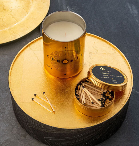Metallic gold glass candle with opened match storage lid rests on a gold platform