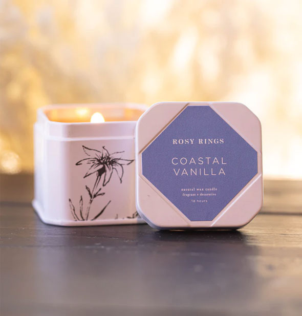 Small white lid candle tin with flower illustration on one side features a debossed lid with blue label that says, "Rosy Rings Coastal Vanilla"