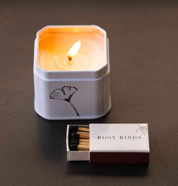 Small white lit tin candle with flower illustration on one side includes a pack of Rosy Rings matches