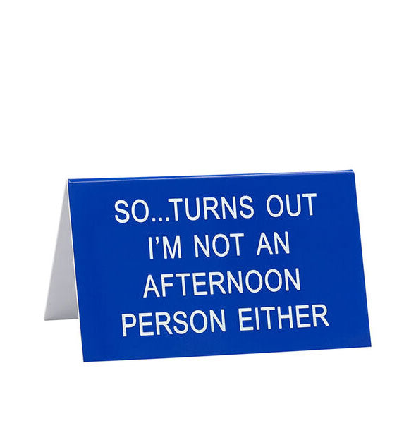 Blue triangular desk sign says, "So...turns out I'm not an afternoon person either" in white lettering
