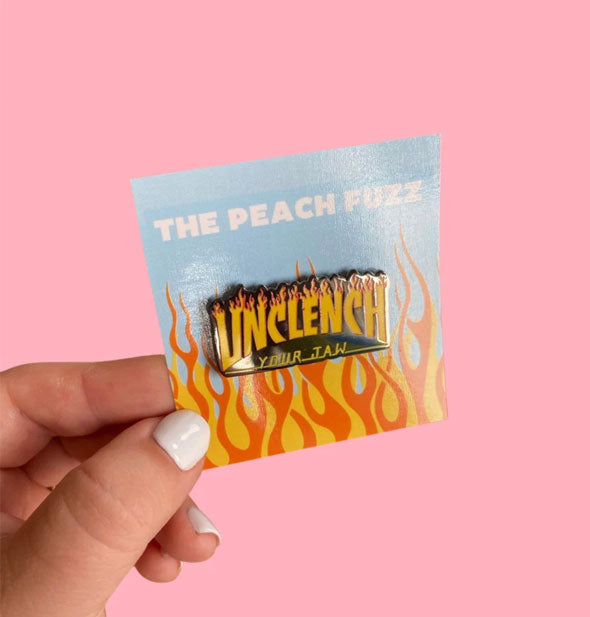 Model's hand holds a black and orange Unclench Your Jaw enamel pin with flame design on a product card by The Peach Fuzz against a pink background