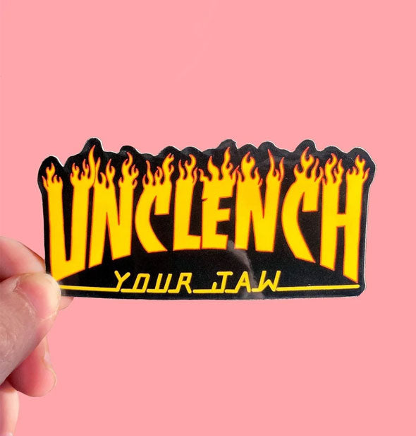 Model's hand holds a sticker with flaming font that says, "Unclench Your Jaw"