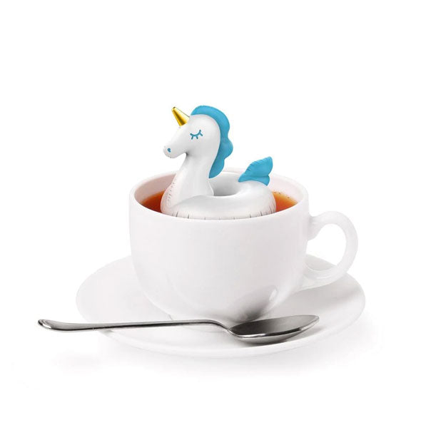 A tea diffuser that resembles an inflatable pool toy floats on the surface of tea inside a white mug resting on a saucer with spoon