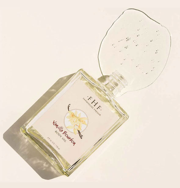 Bottle of FarmHouse Fresh Vanilla Bourbon Body Oil on its side spills oil out onto a light colored surface