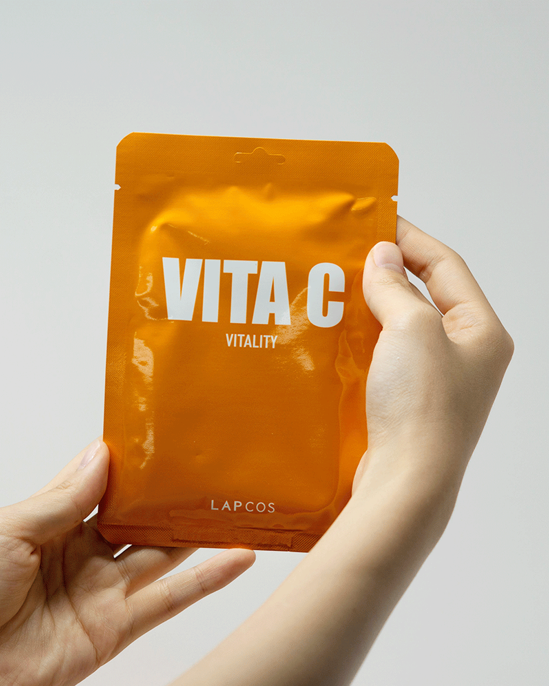 Model's hands open a LAPCOS Vita C Vitality sheet mask pack and unfold the mask inside