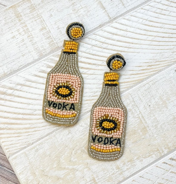 Pair of gray, yellow, pink, and dark blue "Vodka" bottle dangle drop earrings on a weathered white wooden surface