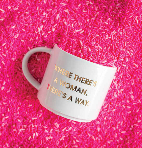 White mug with metallic gold "Where There's a Woman, There's a Way" imprint rests on a backdrop of pink sprinkles