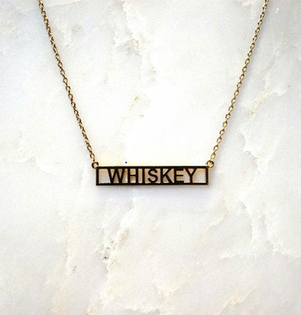 Gold bar necklace on white marble surface says, "Whiskey"