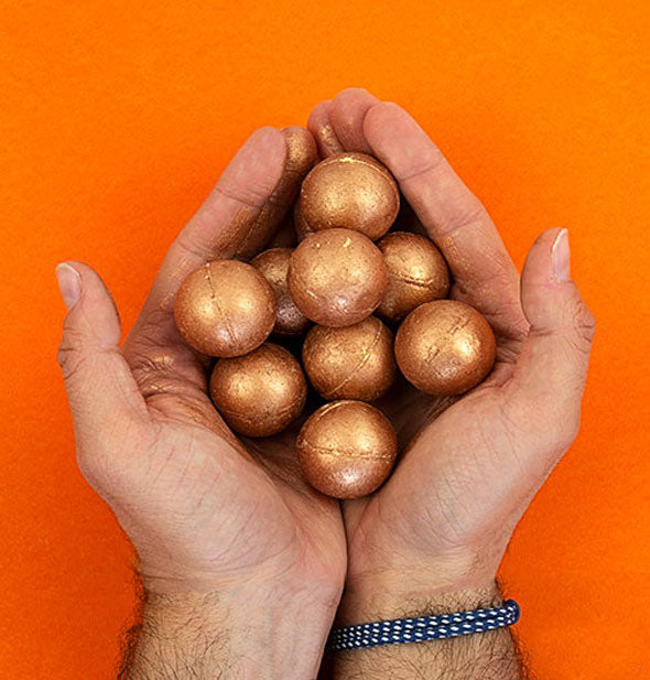 Model's hands cup a handful of shiny copper-colored bath bombs against an orange backdrop