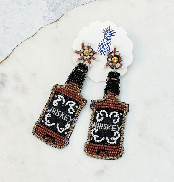Pair of black, brown, white, and gold beaded "Whiskey" bottle dangle drop earrings on a round white card with scalloped edge and blue pineapple logo