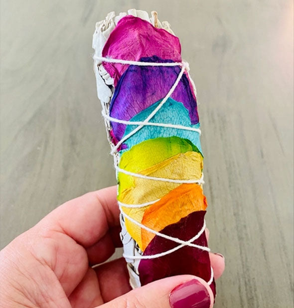 A hand holds a sage smudge stick wrapped with colorful rose petals in rainbow hues