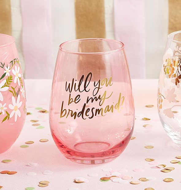 Will You Be My Bridesmaid? wine glass sits on a confetti-covered tabletop between two other decorative stemless wine glasses